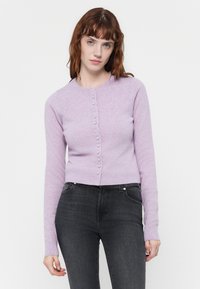 Cárdigan lila con mangas largas, cuello redondo y cierre de botones en la parte delantera. Textura suave, diseño ajustado, combinado con jeans oscuros.