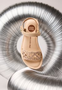 Sandale beige Gios Eppo avec bride avant tressée et boucle à la cheville, centrée sur un fond de tube métallique argenté enroulé.