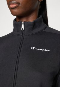 Veste noire à zip en tissu doux avec un col montant, une fermeture éclair argentée et un logo "Champion" blanc sur la poitrine gauche.