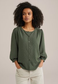 Blusa verde con bottoni, realizzata in tessuto testurizzato, con scollo rotondo e maniche puff a tre quarti, caratterizzata da una vestibilità morbida e motivi a pois discreti.