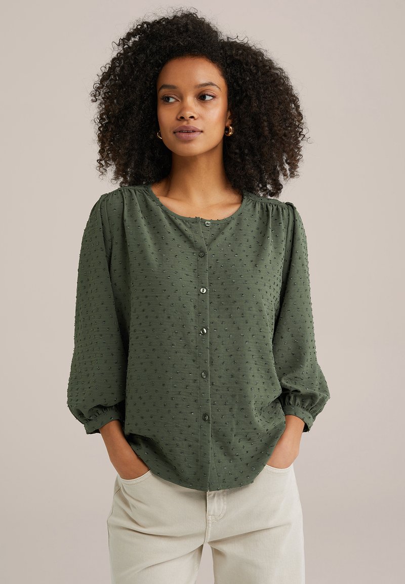 Blusa verde con bottoni, realizzata in tessuto testurizzato, con scollo rotondo e maniche puff a tre quarti, caratterizzata da una vestibilità morbida e motivi a pois discreti.
