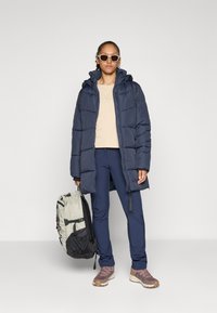 Marine pufferjack met capuchon, beige top, marineblauwe broek en grijze rugzak. Bruine en grijze sneakers. Zonnebril met een lichtgekleurde montuur.