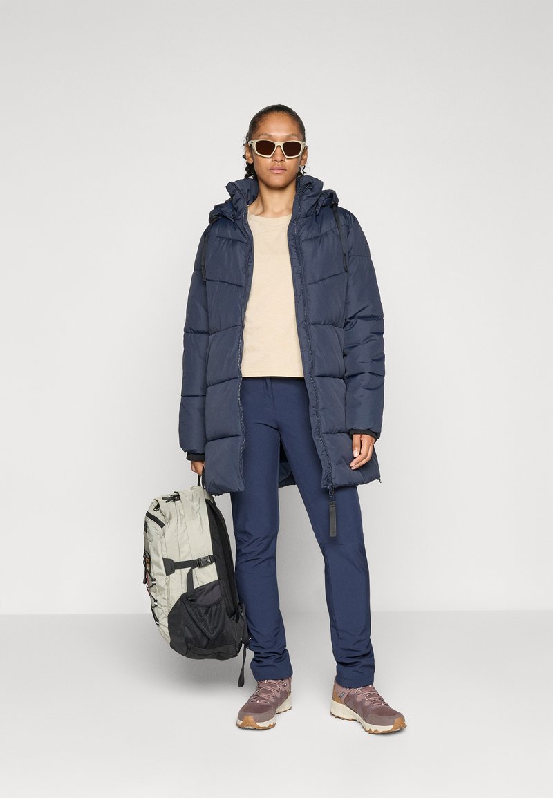 Doudoune marine avec capuche, haut beige, pantalon marine et sac à dos gris. Baskets marron et gris. Lunettes de soleil avec monture claire.