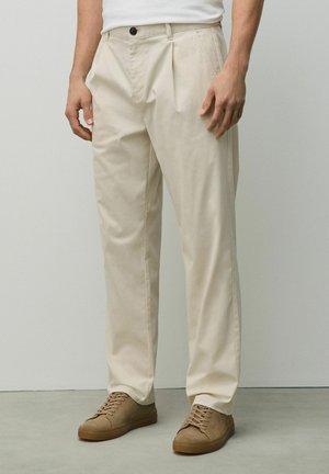 Man wearing beige pleated trousers, white tucked-in shirt, and brown suede lace-up shoes against a plain light background.
