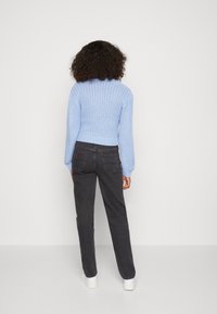 Personne avec des cheveux bouclés portant un pull en tricot bleu clair, un jean noir et des chaussures blanches, se tenant face à un mur neutre et uni.