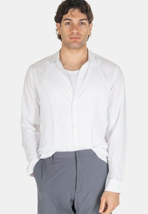 Giovane uomo con capelli scuri e ricci che indossa una camicia bianca abbottonata sopra una canottiera bianca e pantaloni grigi, in piedi con le mani in tasca.