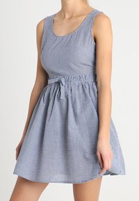 Modré a bílé kostkované šaty s výstřihem do kulata, přiléhavým živůtkem, nařaseným pasem a sukní, která se rozšiřuje. K pasu jsou připevněny mašle.