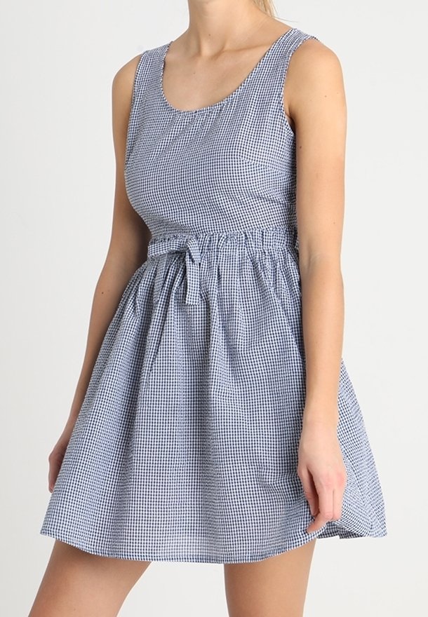 Modré a bílé kostkované šaty s výstřihem do kulata, přiléhavým živůtkem, nařaseným pasem a sukní, která se rozšiřuje. K pasu jsou připevněny mašle.