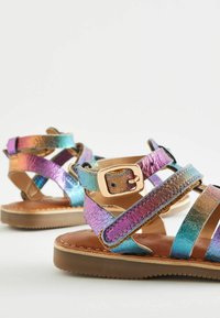 Sandaler med remmar som är iriserande i flera färger, en slät brun innersula och en guldspänne som accent på anklaren.