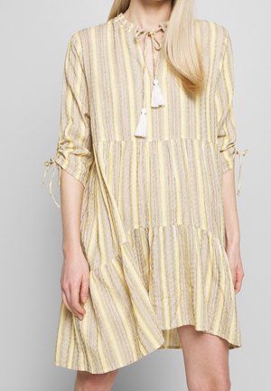 Nainen, jolla on yllään löysä polvipituinen mekko, jossa on pystysuorat keltaiset, beige ja valkoiset raidat, solmittava tupsukaulus ja rypytetyt 3/4-hihat rusetilla.