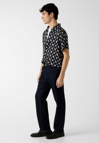 Chemise noire à manches courtes avec des motifs géométriques blancs, assortie à un jean droit bleu foncé et des mocassins noirs texturés.