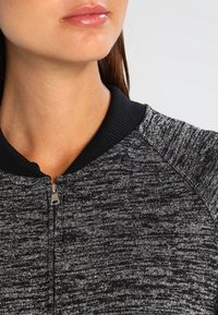 Grijze zip-up sweater met textuurstof, zwarte geribbelde kraag en rits aan de voorkant. Close-up weergave die de materialen en ontwerpdetails accentueert.