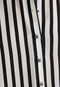 Blusa a rayas en blanco y negro con una tapeta de botones y detalles decorativos. Confeccionada en una tela suave y ligera con una textura agradable.
