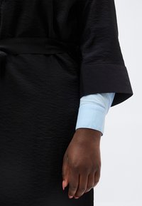 Σκούροχρωμο χέρι με μανσέτα ανοιχτού γαλάζιου πουκάμισου να διακρίνεται κάτω από φαρδύ μαύρο υφασμάτινο μανίκι και ζώνη σε μαύρο ένδυμα.