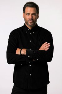 Chemise en velours côtelé noire avec col boutonné, boutons blancs et poche poitrine ; le mannequin a les bras croisés, mettant en valeur l'ajustement et la texture de la chemise.
