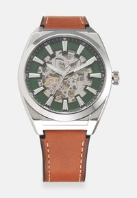 Montre en acier inoxydable argenté avec un cadran squelettique transparent, des accents verts, des marqueurs blancs et un bracelet en cuir brun.