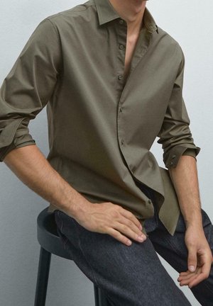 Homme assis sur un tabouret noir portant une chemise boutonnée vert olive avec les manches retroussées et un pantalon gris foncé sur un fond uni.
