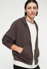 Brun hettegenser med glidelås, store lommer, ribbestrikkede mansjetter og foret hette. Bortover en hvit skjorte med blå stripet detaljer. Myk stofftekstur.