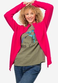 Giacca in pile fucsia con zip frontale, indossata sopra una maglietta verde oliva con una grafica stilizzata di un uccello blu, abbinata a jeans in denim.