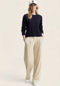 Tengerészkék pulóver, széles bézs nadrág és fehér sportcipő. A modell baseballsapkát visel kék részlettel. Laza szabású, hétköznapi öltözék.