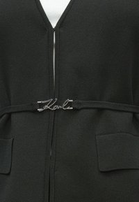 Čierny pletený kardigan s hlbokým výstrihom do V, ozdobený striebornou sponou s logom v páse a dvoma prednými vreckami.