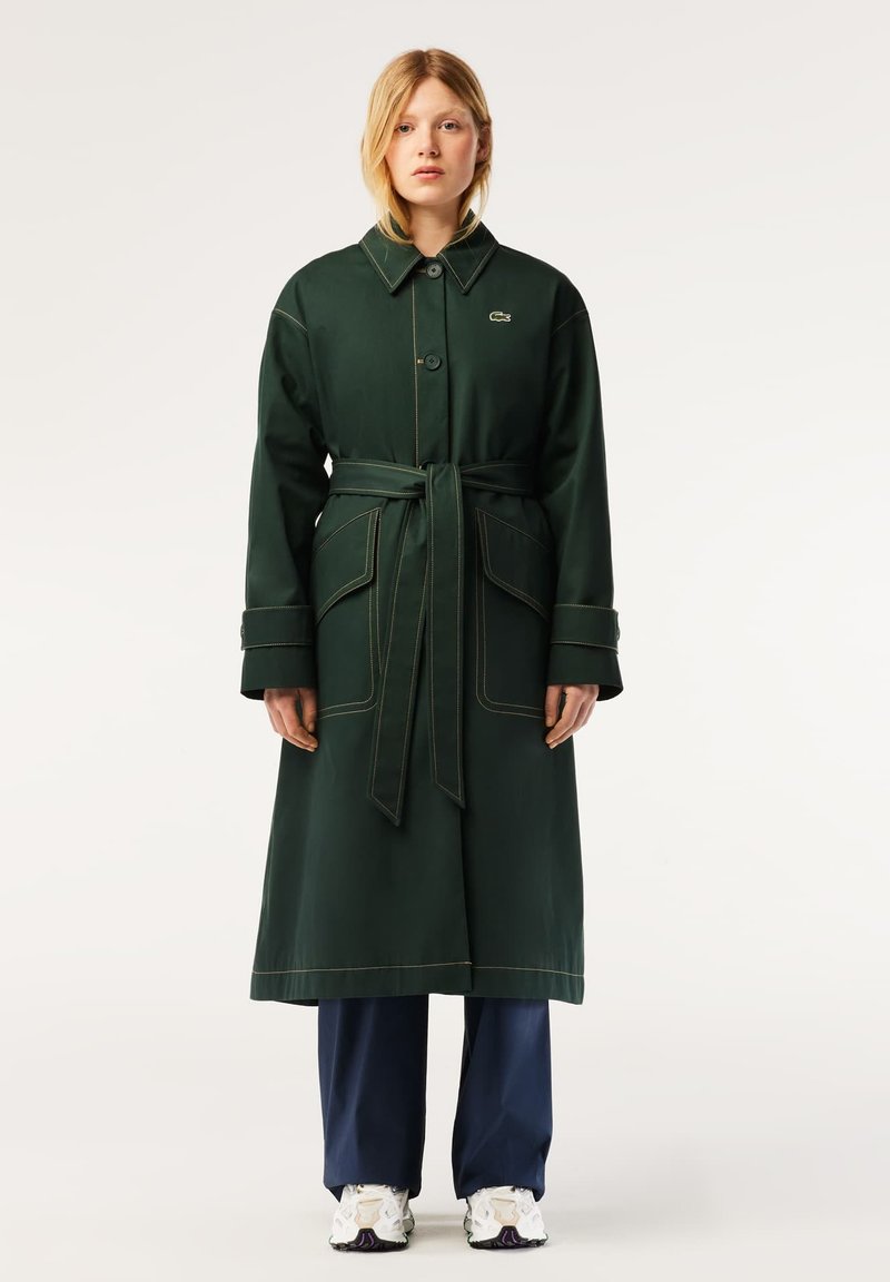 Long manteau vert avec une taille ceinturée, un grand col et des coutures contrastées. Comprend deux poches avant et une fermeture par boutons.