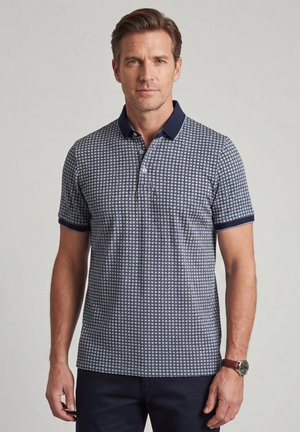 Man die een marineblauw-wit geruit poloshirt draagt met een donker kraagje en manchetten, staand tegen een effen lichte achtergrond.