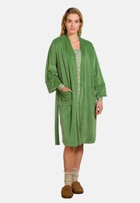 Lang, groen, pluche badjas met wijde mouwen, zijzakken en een striksluiting. Draag deze over een bloemenpatroon jurk, gecombineerd met bruine slippers.