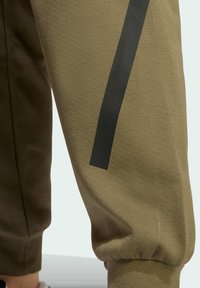 Olivové tepláky s textúrovanou tkaninou, ktoré majú matne čierny vertikálny pruh pozdĺž nohy a elastické manžety na členkoch.