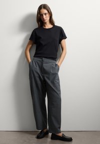 Donna in piedi contro un muro bianco, che indossa una t-shirt nera con maniche arrotolate, pantaloni grigi a gamba larga con tasche e scarpe nere slip-on.