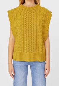 Pull sans manches en tricot jaune moutarde avec un motif torsadé texturé et un ourlet côtelé, associé à un jean bleu clair.