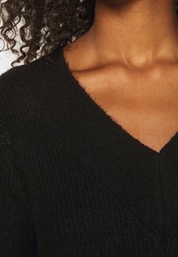 Pull en maille noire avec un col en V profond et une texture côtelée. Des fibres lâches visibles renforcent la qualité tactile.