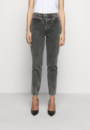 Kobieta w czarnych, wyblakłych jeansach z wysokim stanem i czarnych szpilkach w szpic, stojąca ze swobodnie opuszczonymi rękami wzdłuż ciała na białym tle.