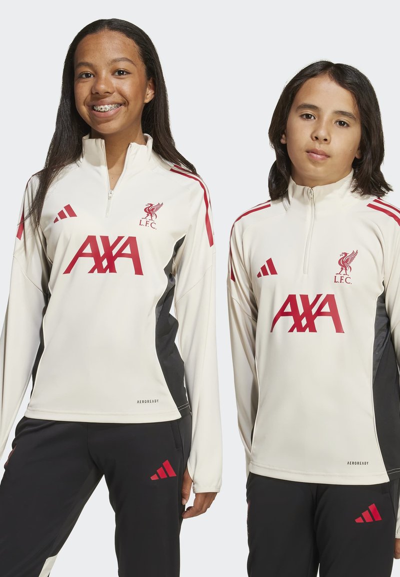 Två barn som bär vita träningströjor från Liverpool FC med röda AXA- och Adidas-logotyper, stående mot en enfärgad bakgrund.