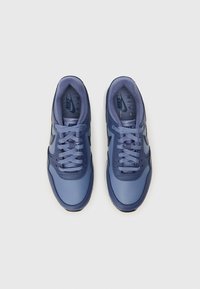 Tênis Nike em diferentes tons de azul, com um cabedal sintético suave, atacadores redondos e uma sola texturizada. Logotipo visível na lingueta.