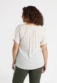 Blusa bianca a maniche corte con ricamo dorato, scollatura arricciata e dettaglio con laccetti regolabili all'orlo, abbinata a pantaloni verde oliva.