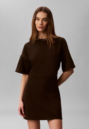 Frau mit glatten braunen Haaren, die ein dunkelbraunes kurzärmeliges Kleid mit Calvin Klein Logo am Kragen trägt, steht vor einem schlichten grauen Hintergrund.