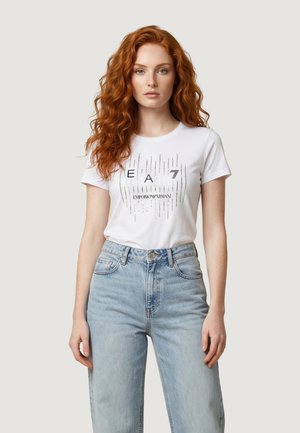 Femme aux cheveux longs, bouclés et roux, portant un t-shirt blanc EA7 Emporio Armani et un jean taille haute bleu clair, posant devant un fond uni.