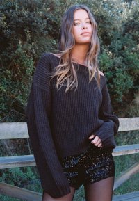 Svart ribbad tröja med överdimensionerade ärmar, kombinerad med glänsande svarta paljettshorts. Naturlig grönska i bakgrunden.