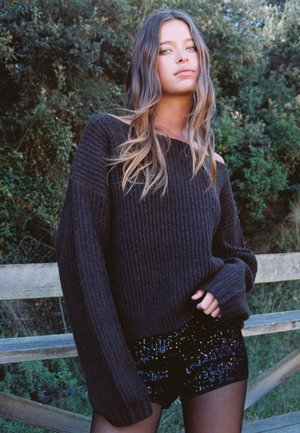 Zwarte geribbelde trui met oversized mouwen, gecombineerd met glanzende zwarte pailletten shorts. Natuurlijke groene achtergrond.