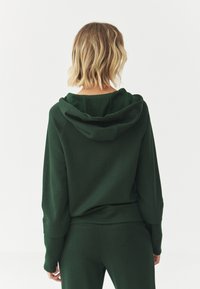 Sudadera verde con un corte relajado, que presenta una gran capucha, puños acanalados y una textura suave. Combinada con pantalones verdes a juego.