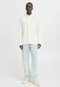Camisa blanca de manga larga con cuello tipo banda y un bolsillo en el pecho, combinada con jeans holgados de corte claro y zapatillas beige.