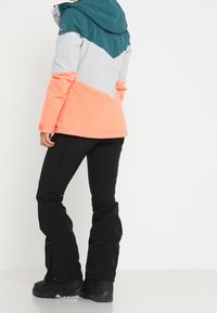 Chaqueta de invierno con bloques de color en tonos verde azulado, gris y coral, con capucha, bolsillos con cremallera y puños ajustables. Combinada con pantalones negros.