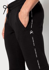 Svarta bomullssweatpants har en dragsnörning i midjan, sidofickor och Calvin Klein Jeans-logotyp i vitt längs vänster ben.