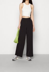 Kvinna som bär en vit kort top, svarta byxor med vida ben, silverfärgade sneakers och håller en grön handväska vid sidan.