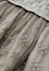 Tissu gris clair avec des motifs floraux brodés et des plis texturés. Le haut présente une bande texturée, légèrement brillante.