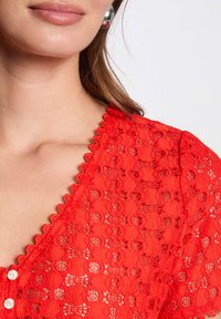 Blouse en dentelle rouge avec un col festonné, présentant des motifs circulaires et deux boutons métalliques. Texture lisse ; design ajusté.