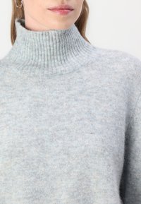 Pull à col roulé bleu clair, côtelé, fabriqué en tissu doux et texturé. Il présente une coupe décontractée et est montré de la poitrine vers le haut.
