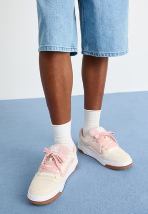 Valged ja roosad tossud kummist tallaga, millel on nubukmaterjal ja nööridega disain. Kantud valgete sokkide ja teksashortsidega.