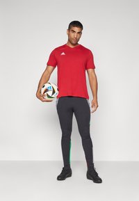 Camiseta atlética roja de manga corta, pantalones ajustados negros con rayas laterales verdes y blancas, sosteniendo un balón de fútbol multicolor, calzado negro.